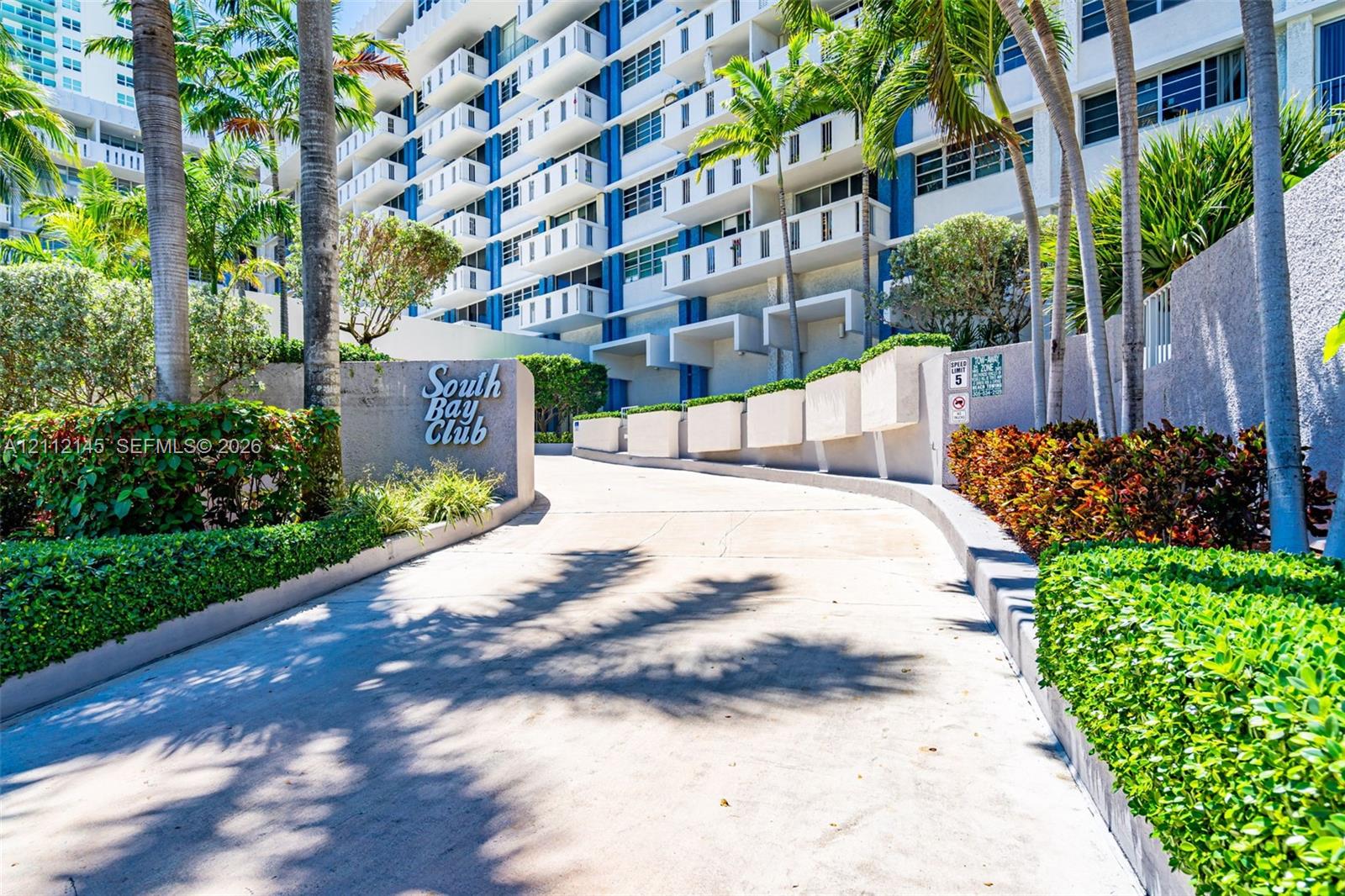 Concrete driveway entrance featuring tropical landscaping, manicured hedges, and multi-story building facade with private balconies Concrete driveway entrance featuring tropical landscaping, manicured hedges, and multi-story building facade with private balconies