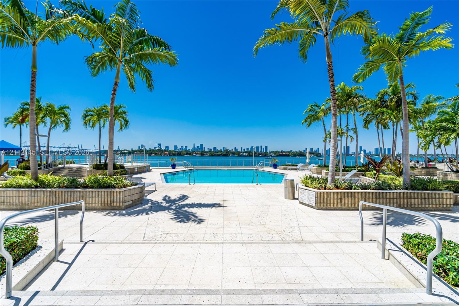 Outdoor pool featuring a paved deck, tiered stone planters, mature palm trees, and waterfront perimeter fencing Outdoor pool featuring a paved deck, tiered stone planters, mature palm trees, and waterfront perimeter fencing