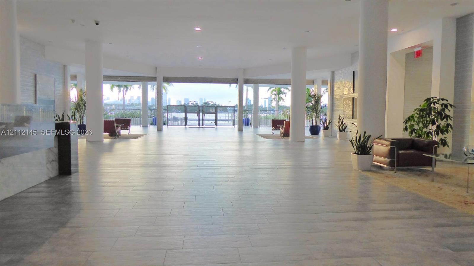 Expansive lobby featuring neutral tile flooring, structural columns, recessed lighting, and a glass-paneled entrance with exterior waterfront scenery Expansive lobby featuring neutral tile flooring, structural columns, recessed lighting, and a glass-paneled entrance with exterior waterfront scenery