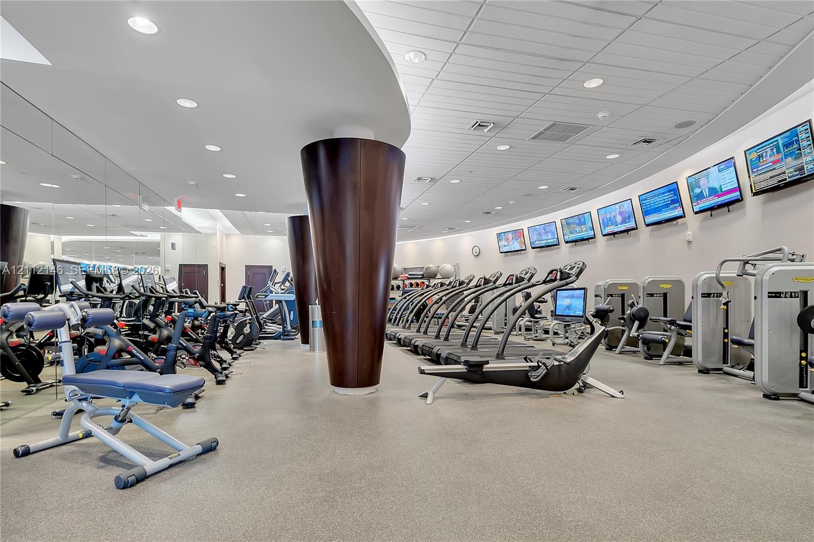 Fitness center featuring a curved wall design, mirrored walls, recessed ceiling lighting, tapered columns with dark wood-finish accents, and multiple wall-mounted television screens