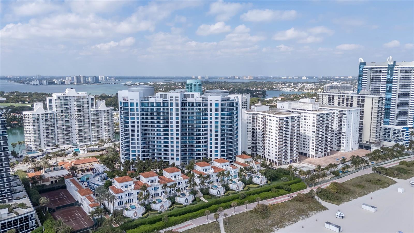 Coastal high-rise complex featuring white sand beach access, terracotta-roofed villas, multiple swimming pools, and tennis courts