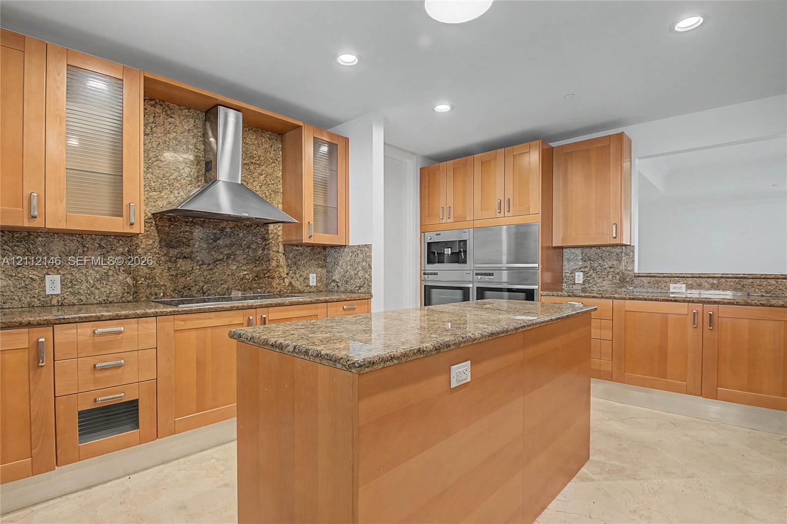Kitchen featuring natural wood cabinetry, stone countertops, a stainless steel chimney-style range hood, integrated wall ovens, and stone tile flooring