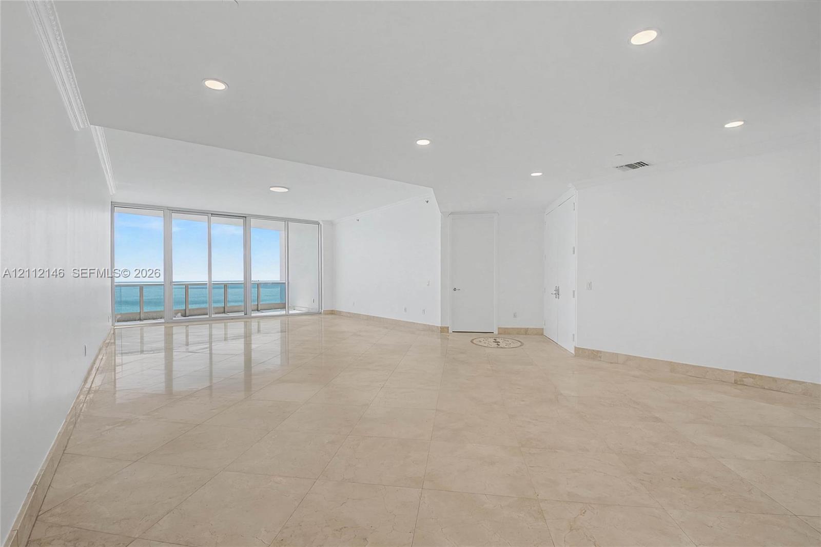 Open-concept interior featuring large-format tile flooring, recessed lighting, floor-to-ceiling glass sliding doors, and a balcony with coastal scenery