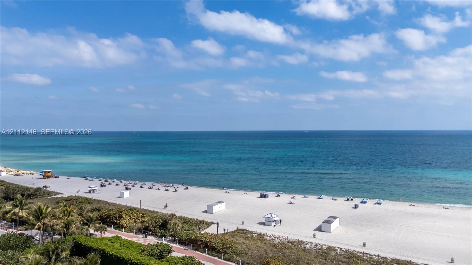 Oceanfront perspective featuring a white sand beach, turquoise water, mature palm trees, and a paved coastal pathway