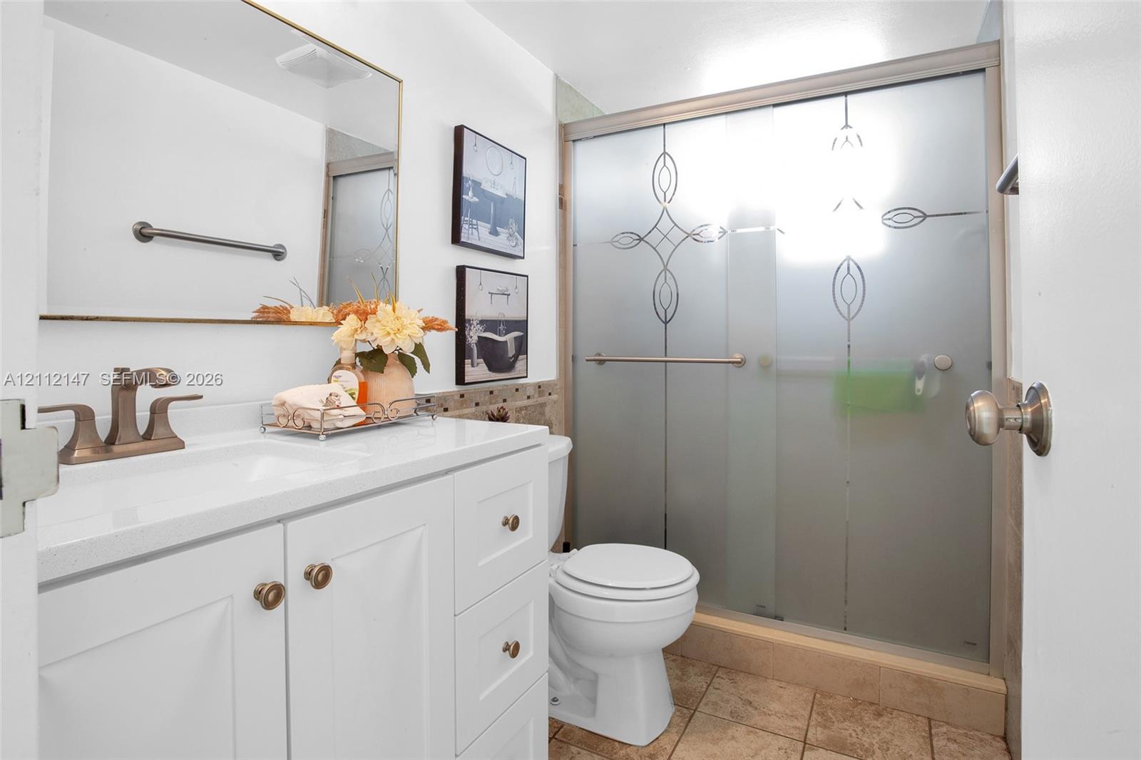 White vanity with a solid-surface countertop and dual-handle faucet, integrated rectangular sink, wall-mounted mirror, and a walk-in shower featuring an etched glass enclosure