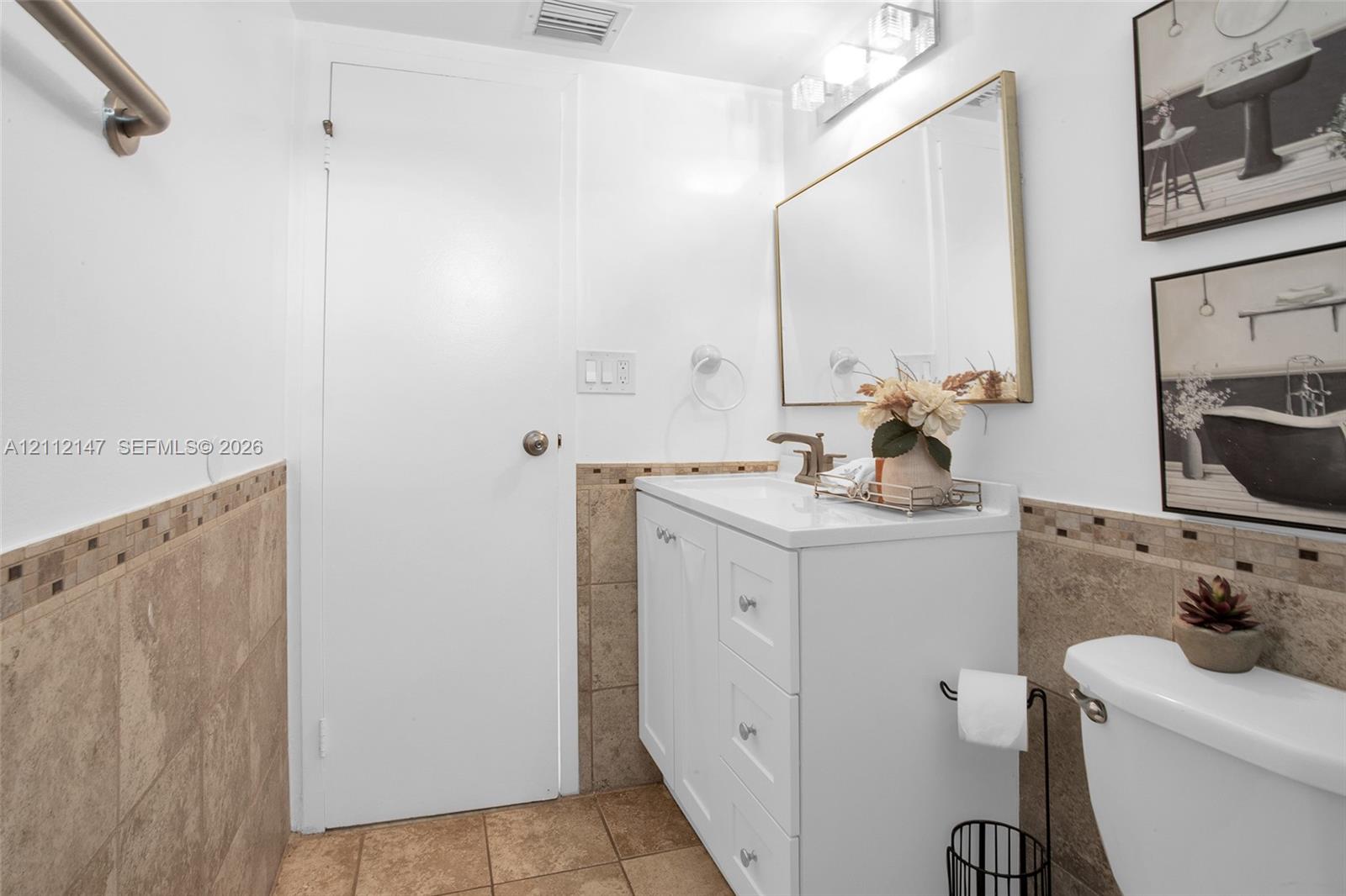 Bathroom featuring a white vanity cabinet with drawers, a rectangular mirror with a metallic frame, neutral-toned wall and floor tile, and a three-light fixture