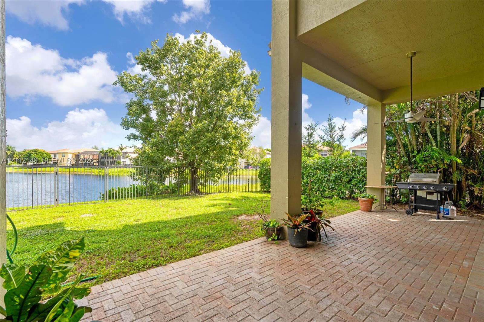 Covered patio featuring brick pavers, ceiling fan, water frontage, perimeter metal fencing, and grass lawn