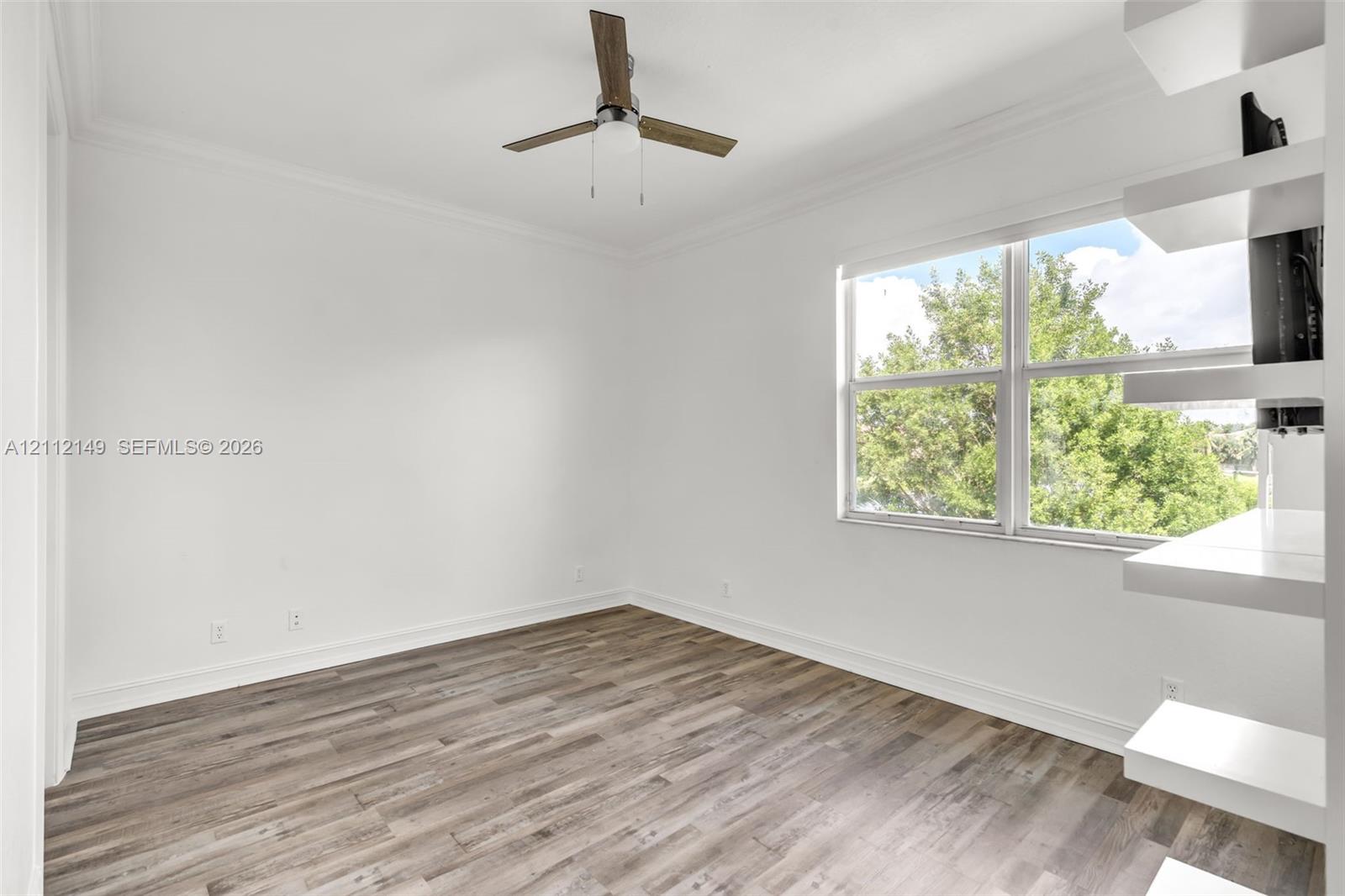 Wood-finish flooring, white built-in shelving, double-pane window, and modern ceiling fan with integrated lighting
