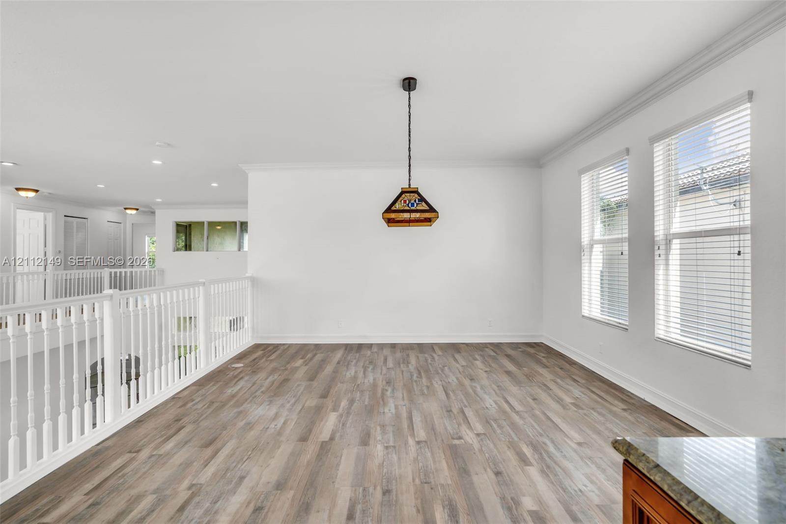 Open space featuring wood-finish flooring, white walls, crown molding, double windows with horizontal blinds, and a suspended geometric pendant light fixture