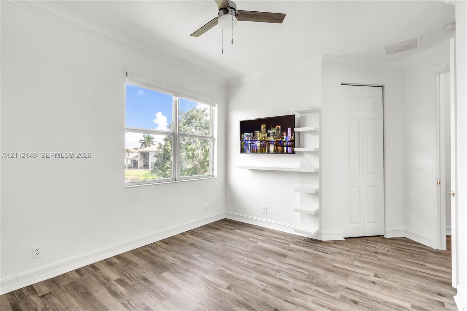 Neutral-toned wood-finish flooring, white built-in shelving, ceiling fan, and multi-pane window