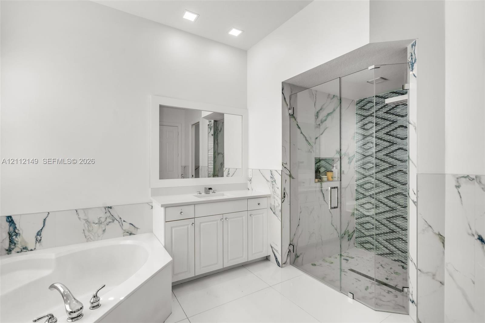White vanity with integrated basin, framed wall mirror, soaking tub with deck-mounted faucet, walk-in shower with glass enclosure, and patterned tile accents