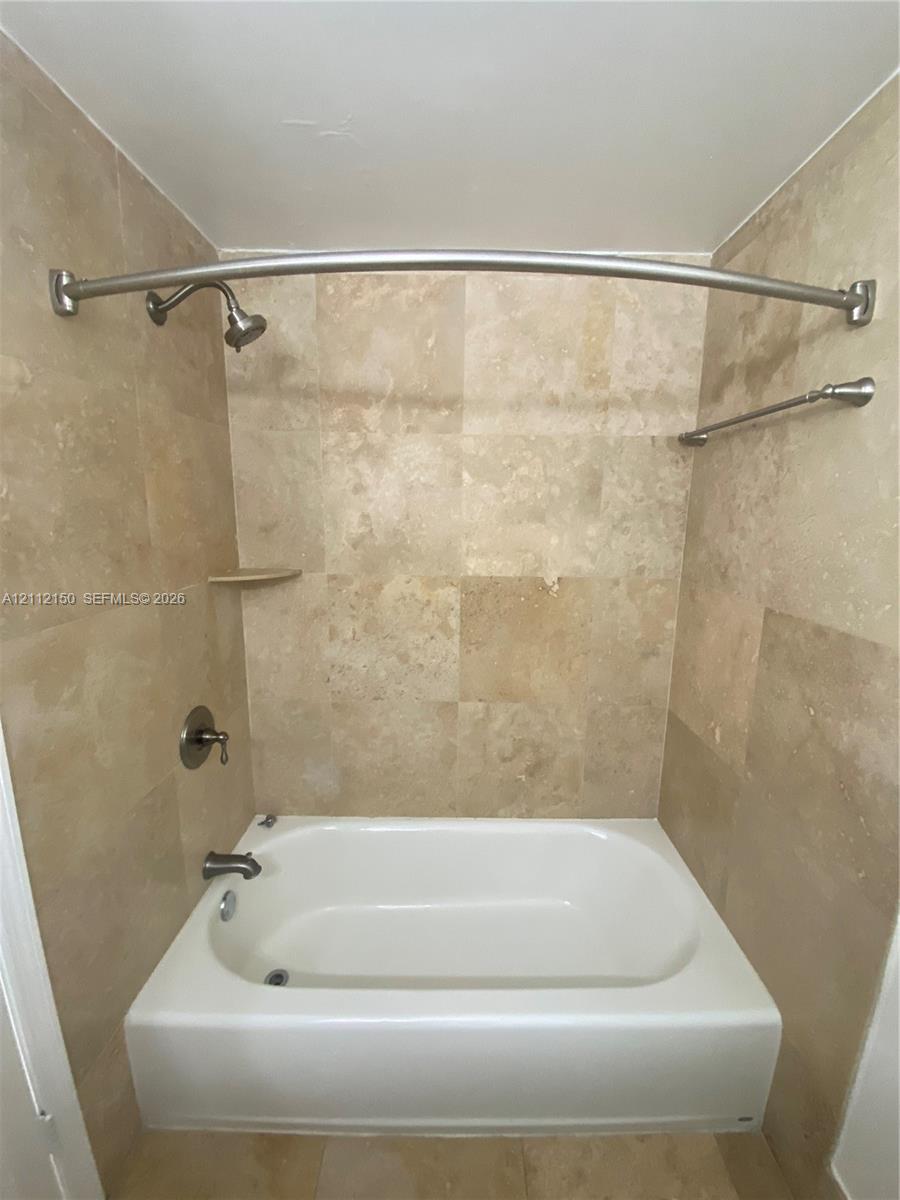 Bathtub enclosure featuring neutral-toned stone wall tile, built-in corner shelf, curved shower rod, and metallic hardware