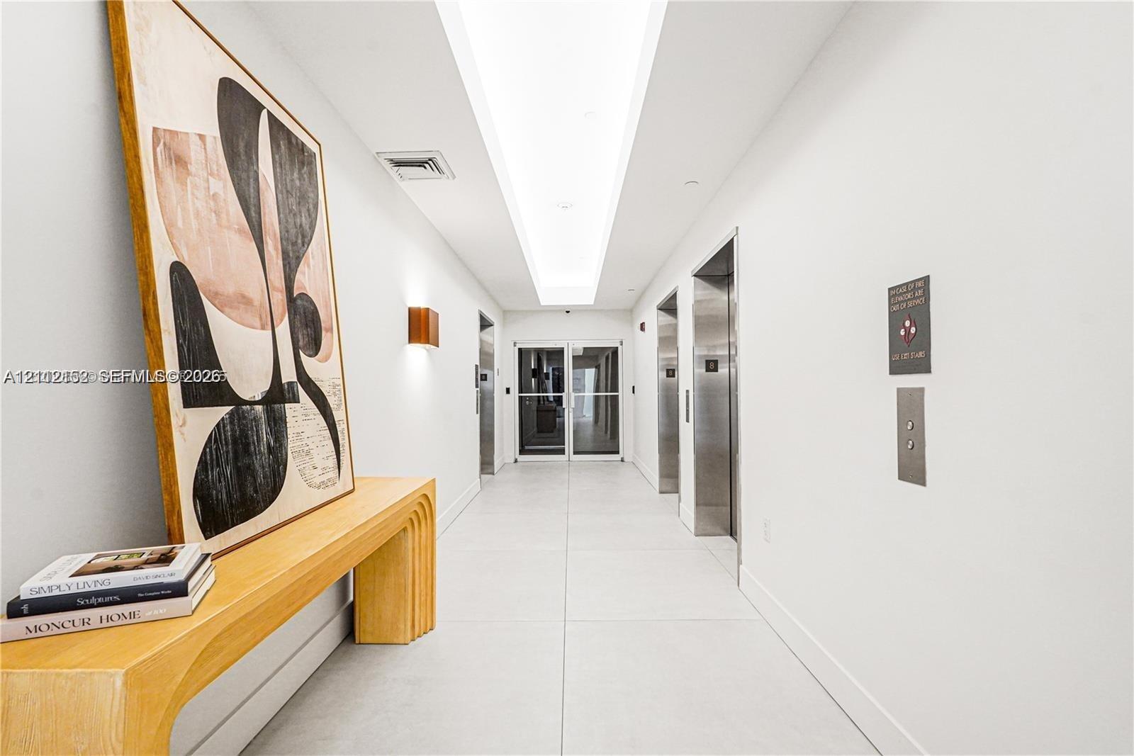 Common area hallway featuring large-format tile flooring, recessed ceiling lighting with a coved center panel, brushed metal elevator doors, and wall-mounted sconces