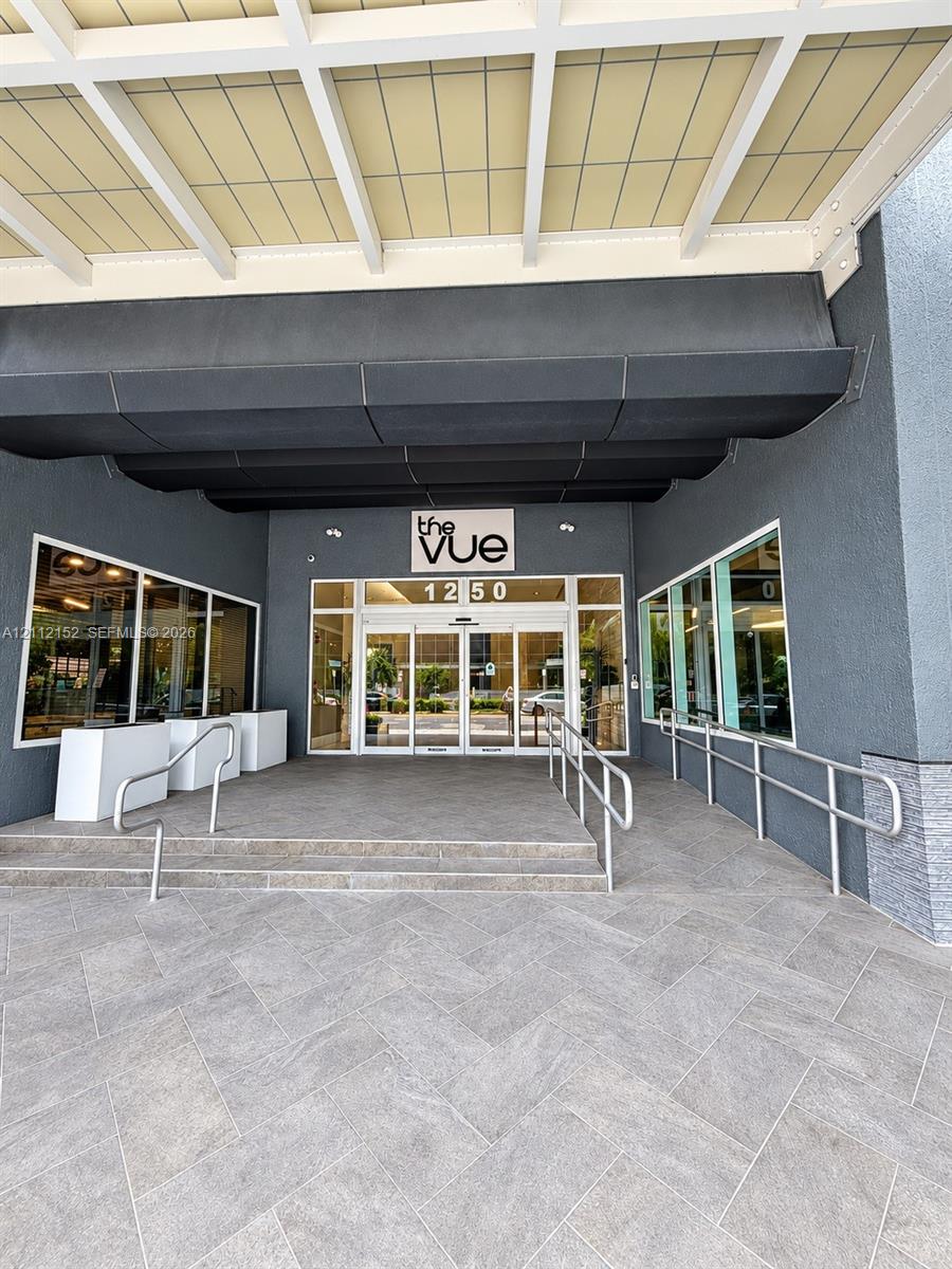 Covered entrance featuring a herringbone-patterned stone floor, glass-paneled automatic doors, metal handrails, and a modern architectural facade