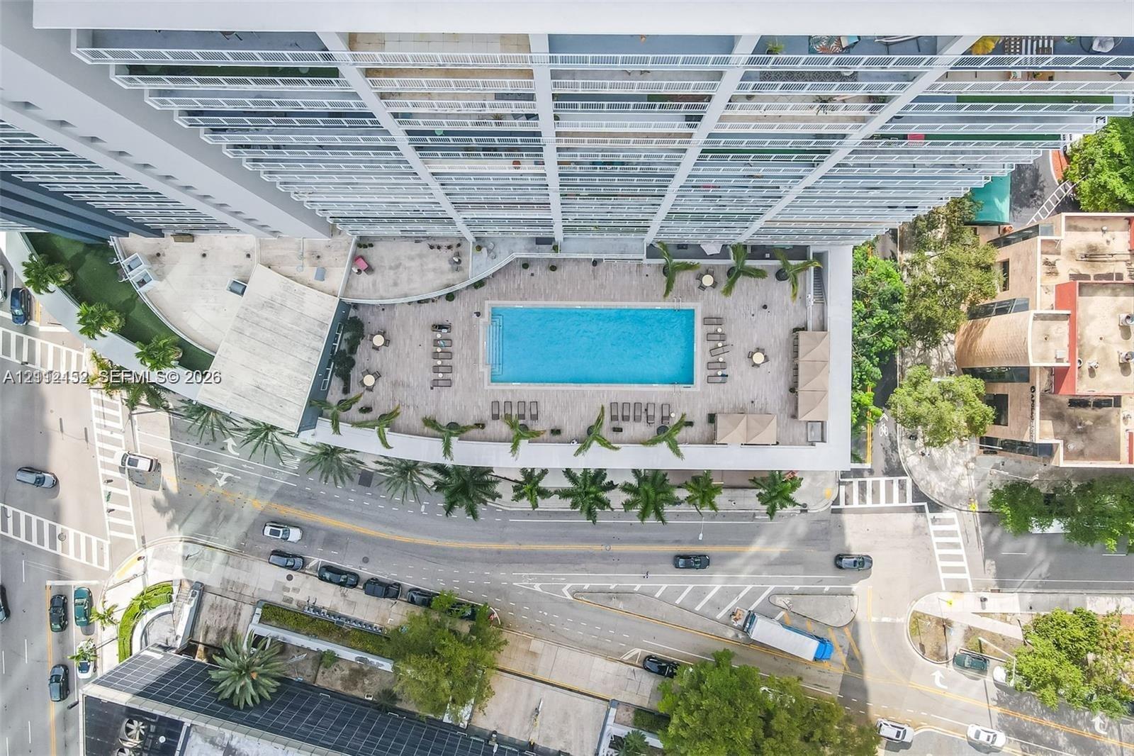Aerial perspective of a high-rise residential structure featuring a central swimming pool, expansive paved pool deck, and integrated palm tree landscaping