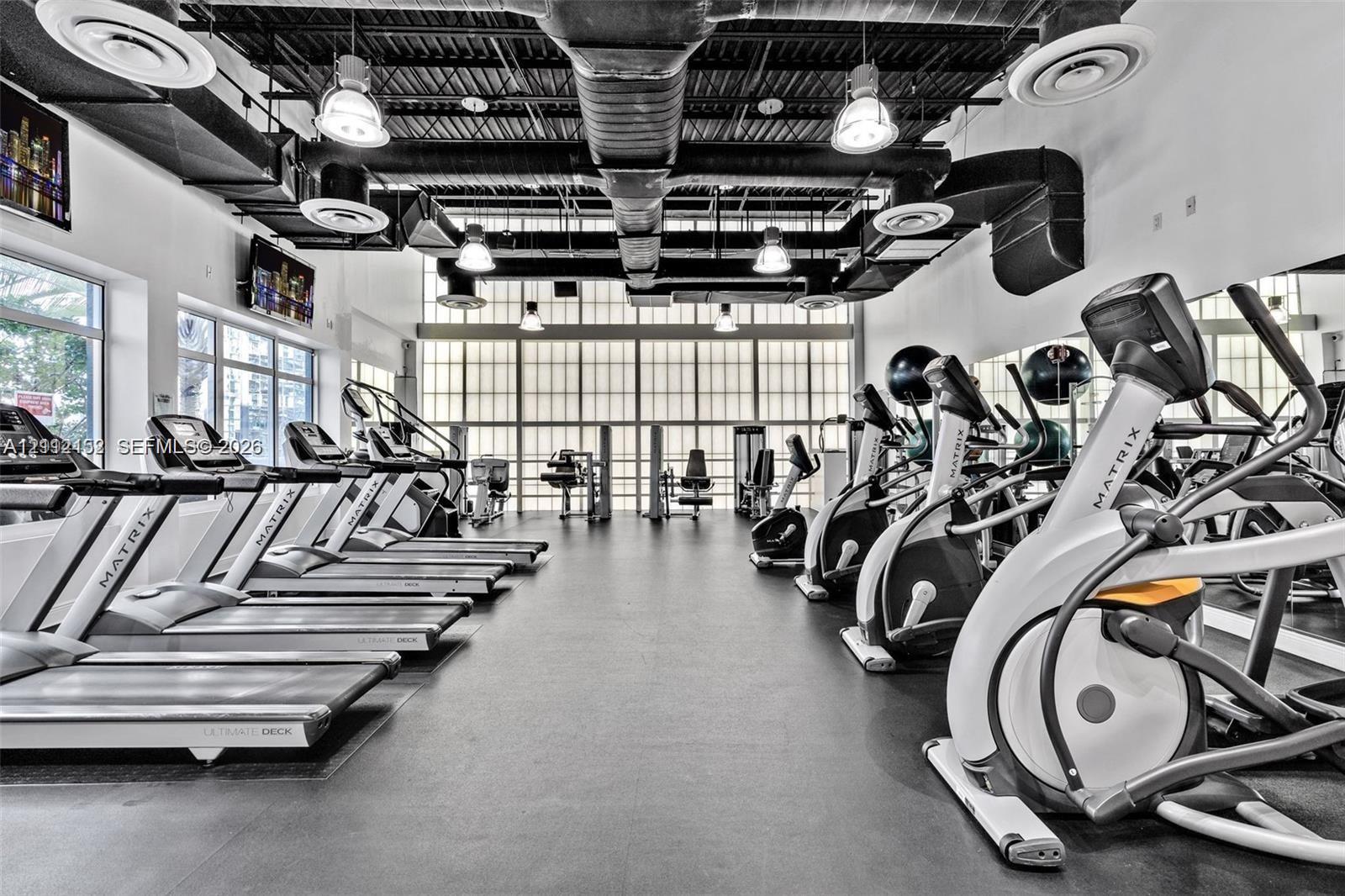 Fitness center featuring high ceilings with exposed industrial-style ductwork, rubberized flooring, expansive grid-patterned windows, and wall-to-wall mirrors
