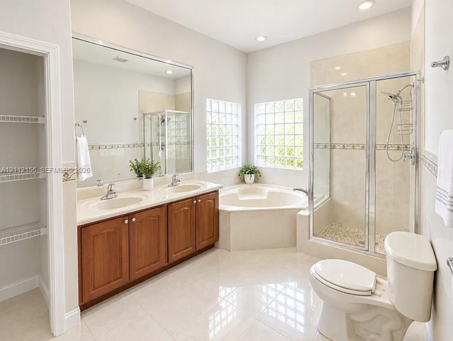 Dual-sink vanity with wood-finish cabinetry, corner soaking tub, glass-enclosed shower, glass block windows, and built-in linen shelving