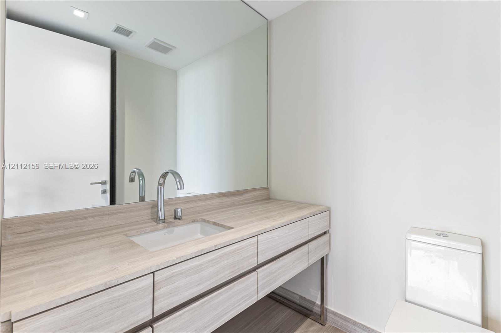Floating vanity featuring wood-finish cabinetry, stone countertop with an integrated rectangular sink, wall-mounted polished chrome faucet, and a large seamless mirror