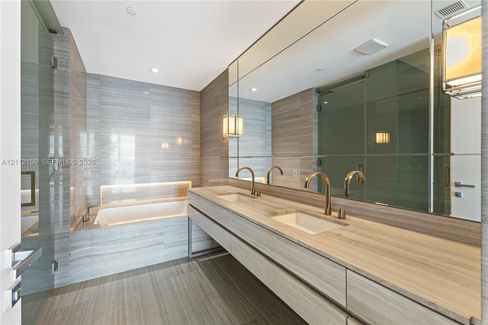 Dual-vanity cabinet featuring stone-topped surfaces, undermount sinks, brass-toned fixtures, floor-to-ceiling tiled walls, and a recessed soaking tub with integrated lighting