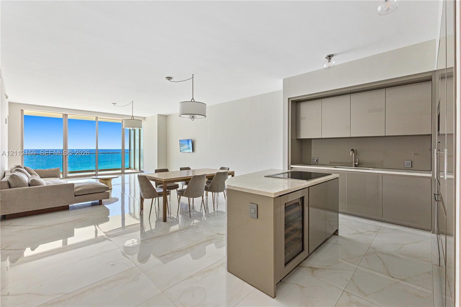 Open-concept interior featuring light-toned flooring, high-gloss flat-panel cabinetry, a stone-topped kitchen island with a built-in wine cooler, recessed lighting, and floor-to-ceiling glass sliding doors Open-concept interior featuring light-toned flooring, high-gloss flat-panel cabinetry, a stone-topped kitchen island with a built-in wine cooler, recessed lighting, and floor-to-ceiling glass sliding doors