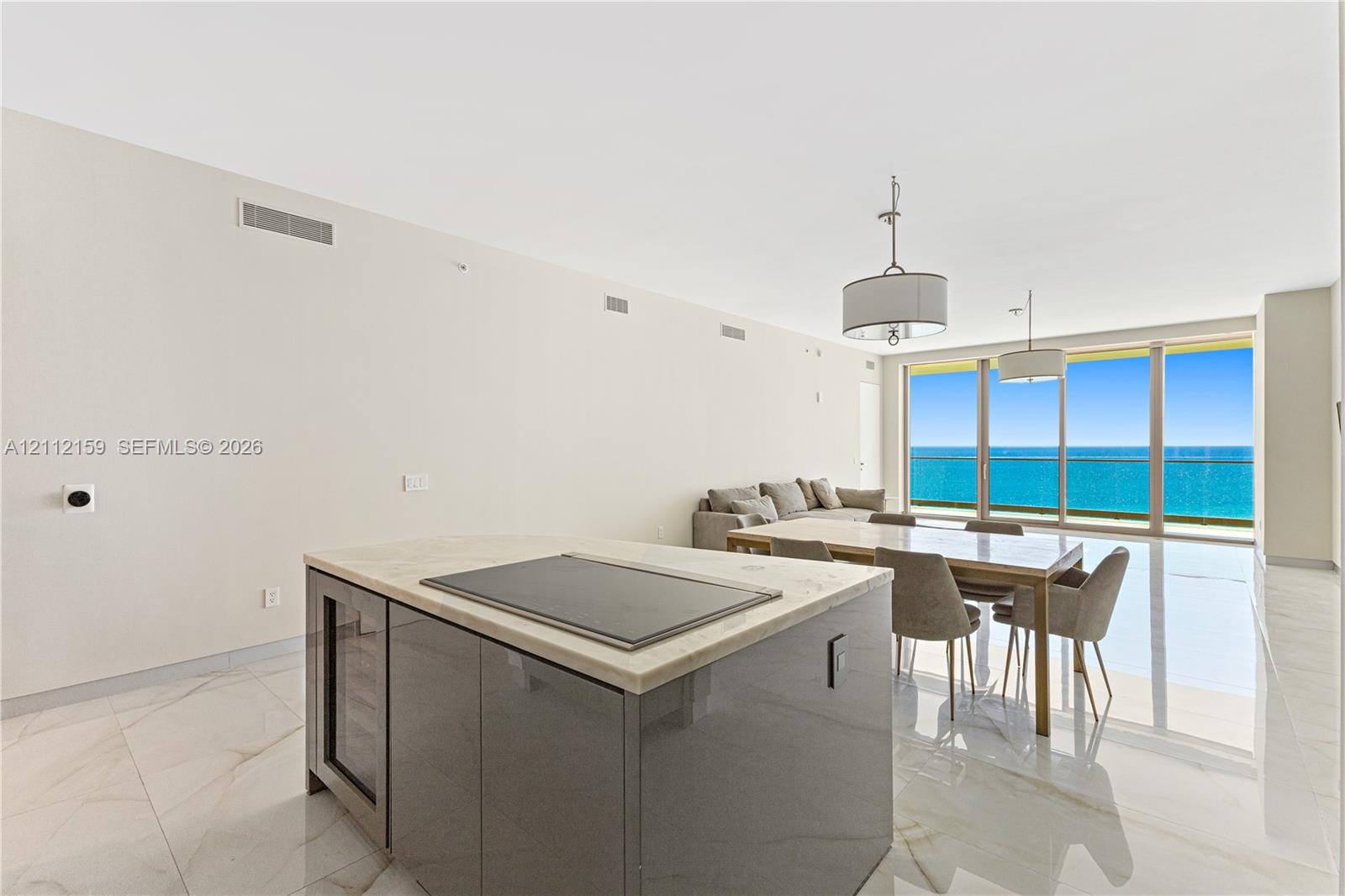 Open-concept interior featuring large-format stone-look flooring, center island with stone countertop and built-in wine refrigeration, dual drum pendant lighting, and floor-to-ceiling glass sliding doors with ocean backdrop Open-concept interior featuring large-format stone-look flooring, center island with stone countertop and built-in wine refrigeration, dual drum pendant lighting, and floor-to-ceiling glass sliding doors with ocean backdrop