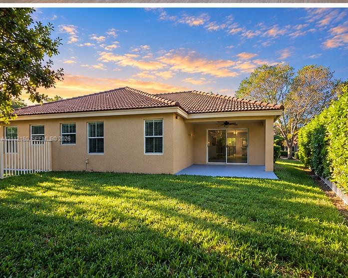 Exterior rear elevation featuring a tile roof, stucco siding, covered patio, and fenced backyard with mature trees