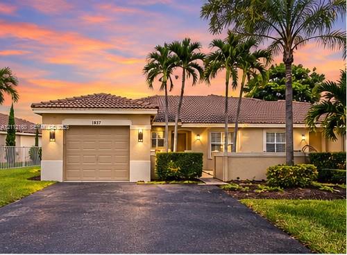 Stucco exterior featuring a tiled roof, attached garage, manicured landscaping, and mature palm trees