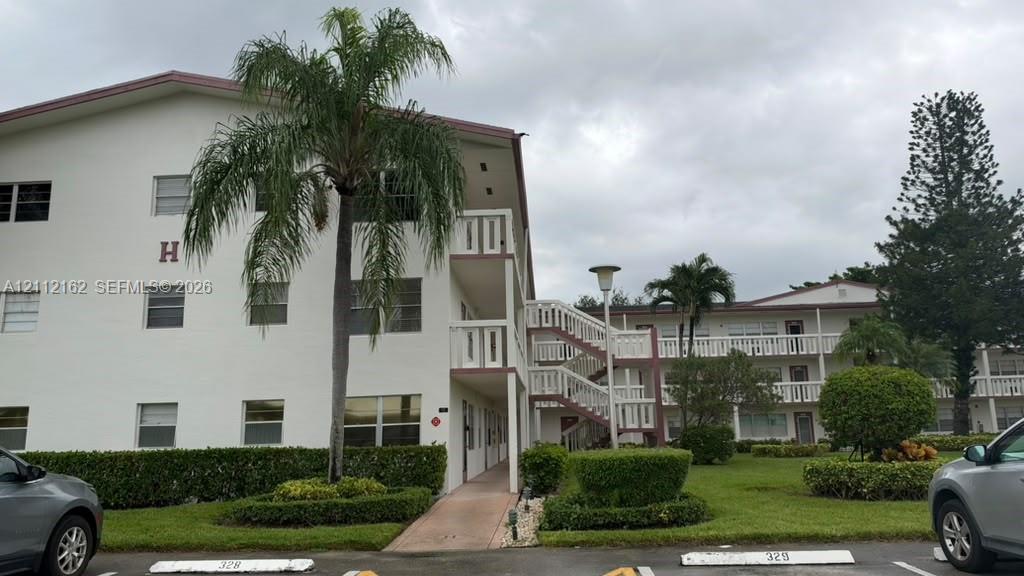 Multi-story building exterior featuring white stucco siding, open-air walkways with balustrade railings, manicured shrubbery, and mature palm trees