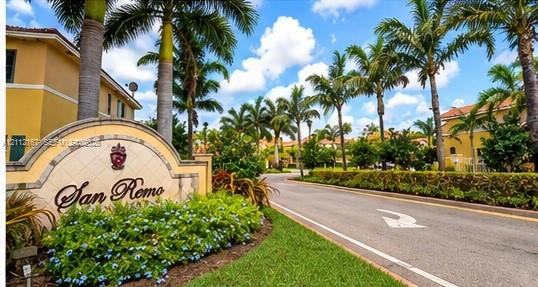 Community entrance featuring a stone-finish monument sign, mature palm trees, manicured landscaping, and asphalt paving