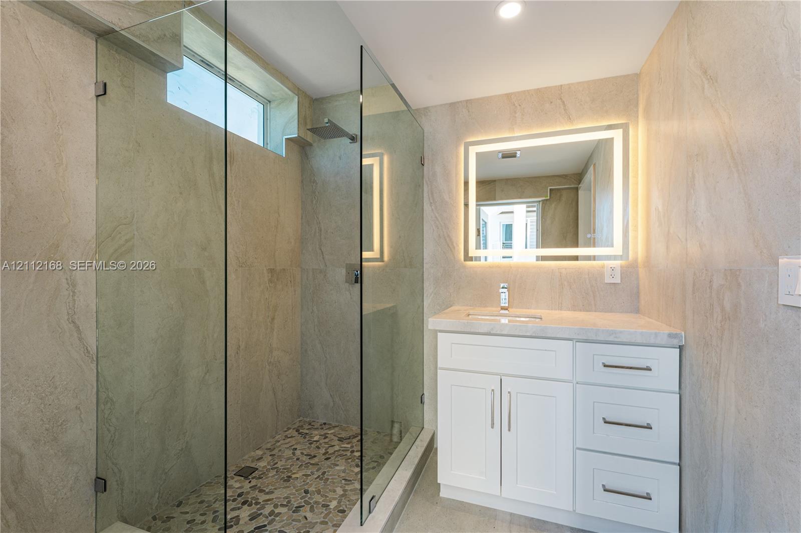 Walk-in shower with a glass enclosure, pebble tile floor, and rainfall showerhead, complemented by a white vanity with a stone countertop, integrated sink, and an illuminated mirror