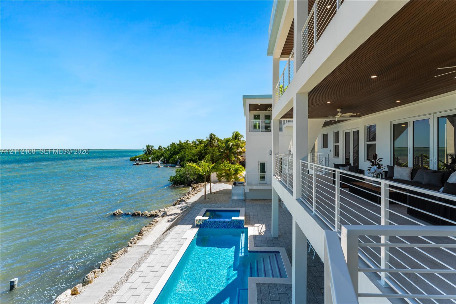 Multi-level waterfront residence featuring a lap pool with an integrated spa, expansive covered balcony with slatted wood-finish ceilings, metal railings, and stone-paver hardscaping