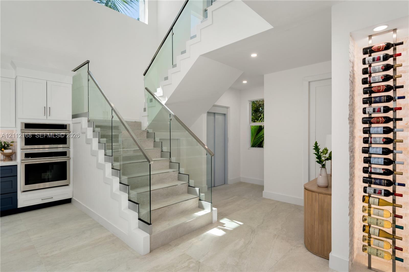 Open-riser staircase featuring a glass-panel railing, built-in stainless steel double wall ovens, recessed wine storage rack, and light-toned tile flooring