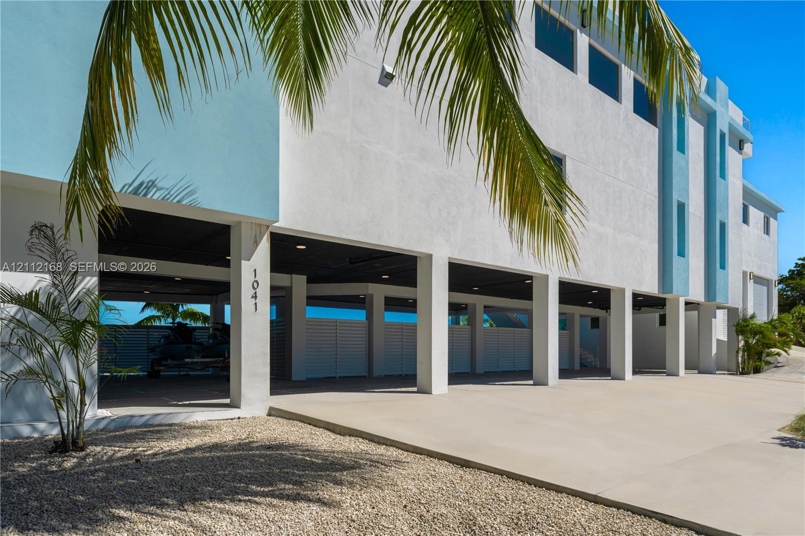 Modern multi-level exterior featuring white stucco finish, covered parking area, concrete driveway, horizontal slat privacy fencing, and recessed lighting