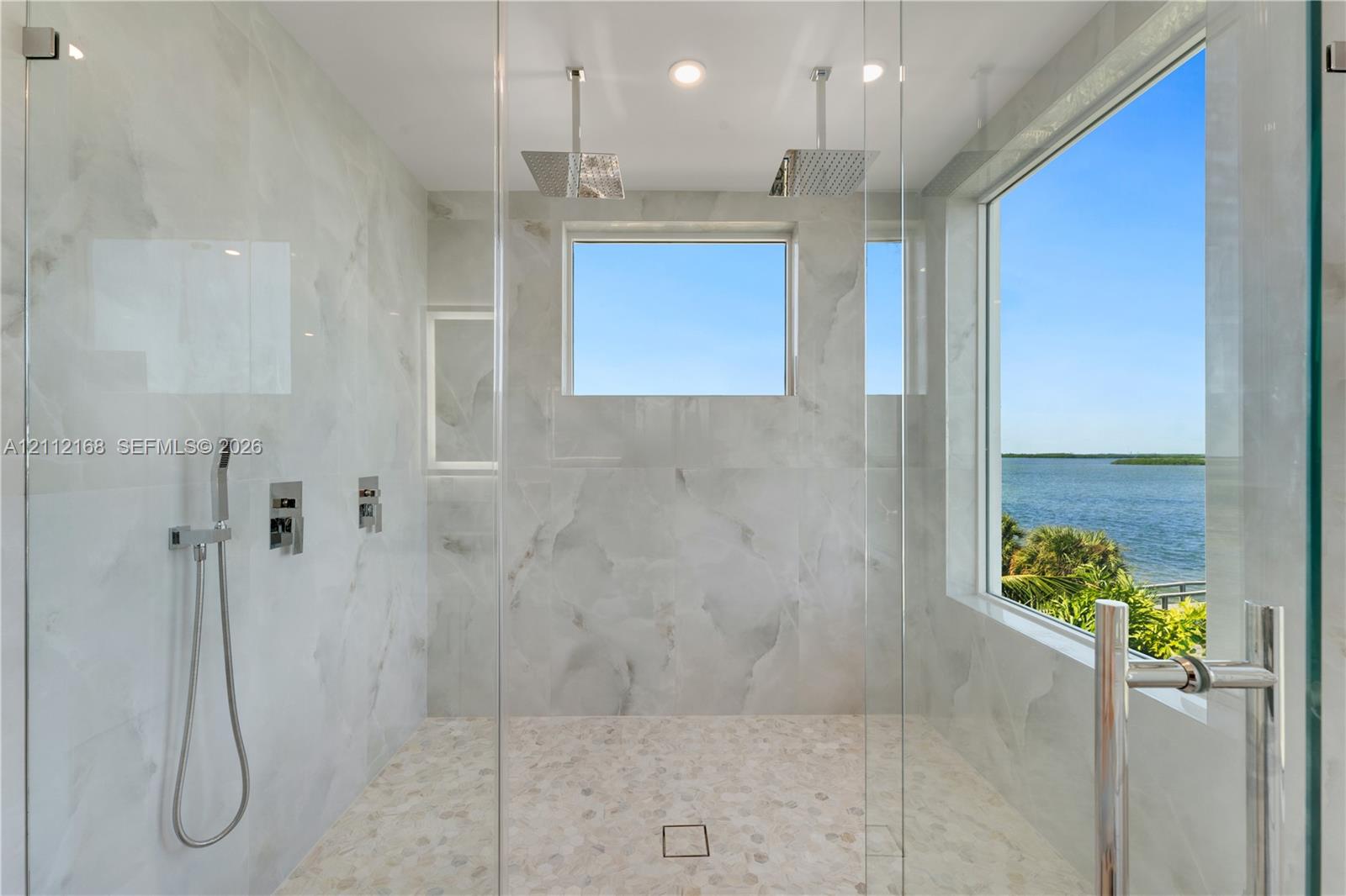 Walk-in shower featuring floor-to-ceiling stone-patterned tile, dual ceiling-mounted rainfall showerheads, a handheld sprayer, a glass enclosure, and a large window