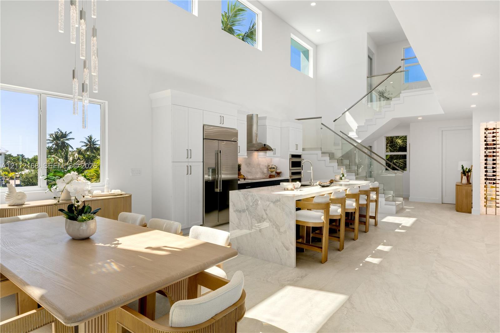 Open-concept kitchen featuring an expansive stone-topped waterfall island, custom white cabinetry, stainless steel professional-grade appliances, floating staircase with glass railings, and double-height ceilings with transom windows