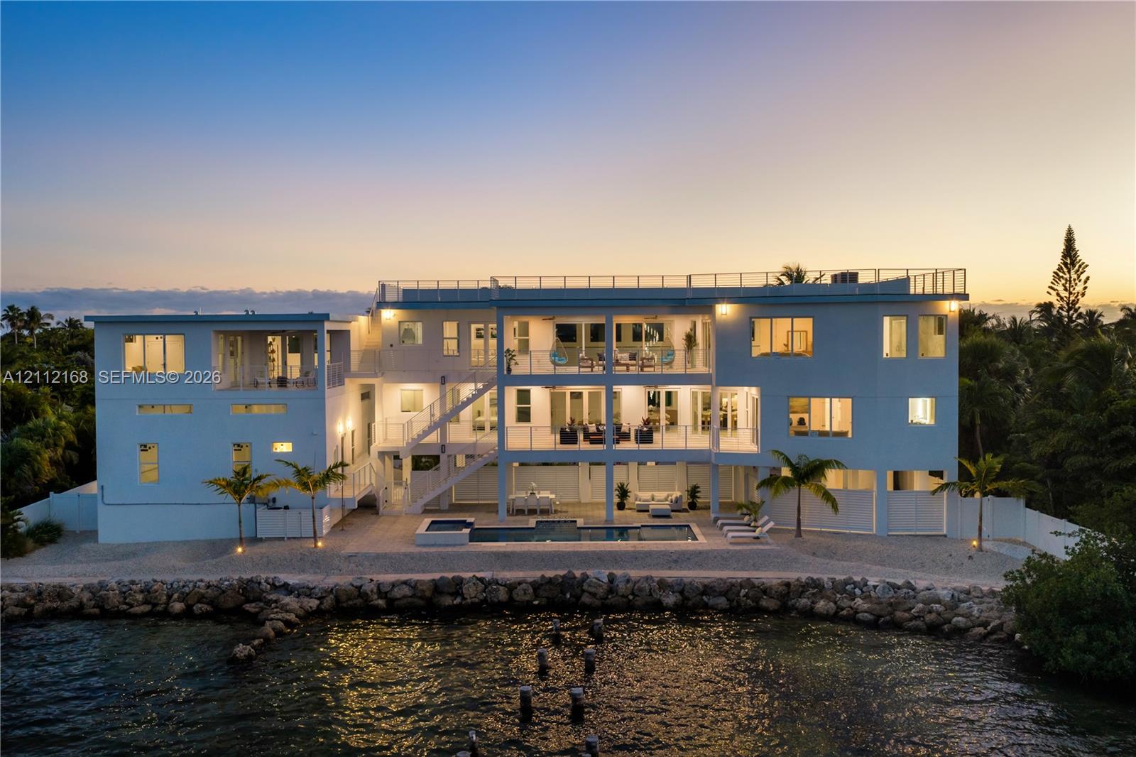 Waterfront multi-story residence featuring a private swimming pool, expansive stacked balconies with metal railings, a rooftop terrace, and a stone-lined bulkhead