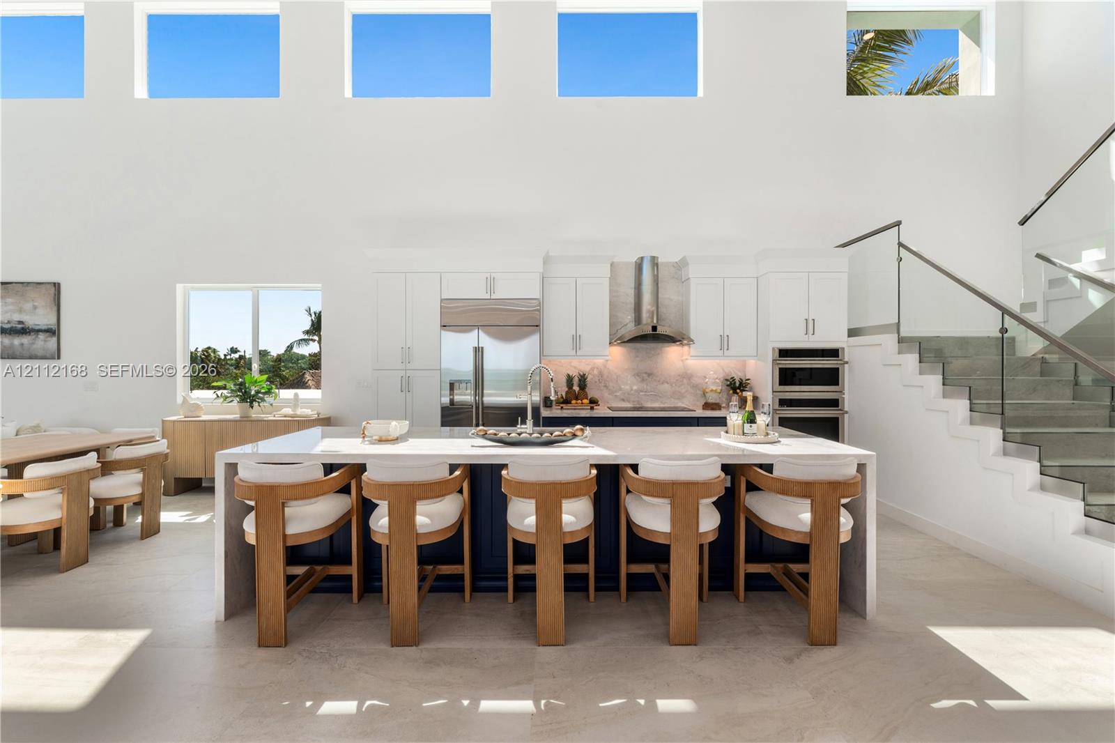 Double-height ceilings with transom windows, large center island with waterfall countertop, stainless steel kitchen appliances, and glass-paneled staircase railing