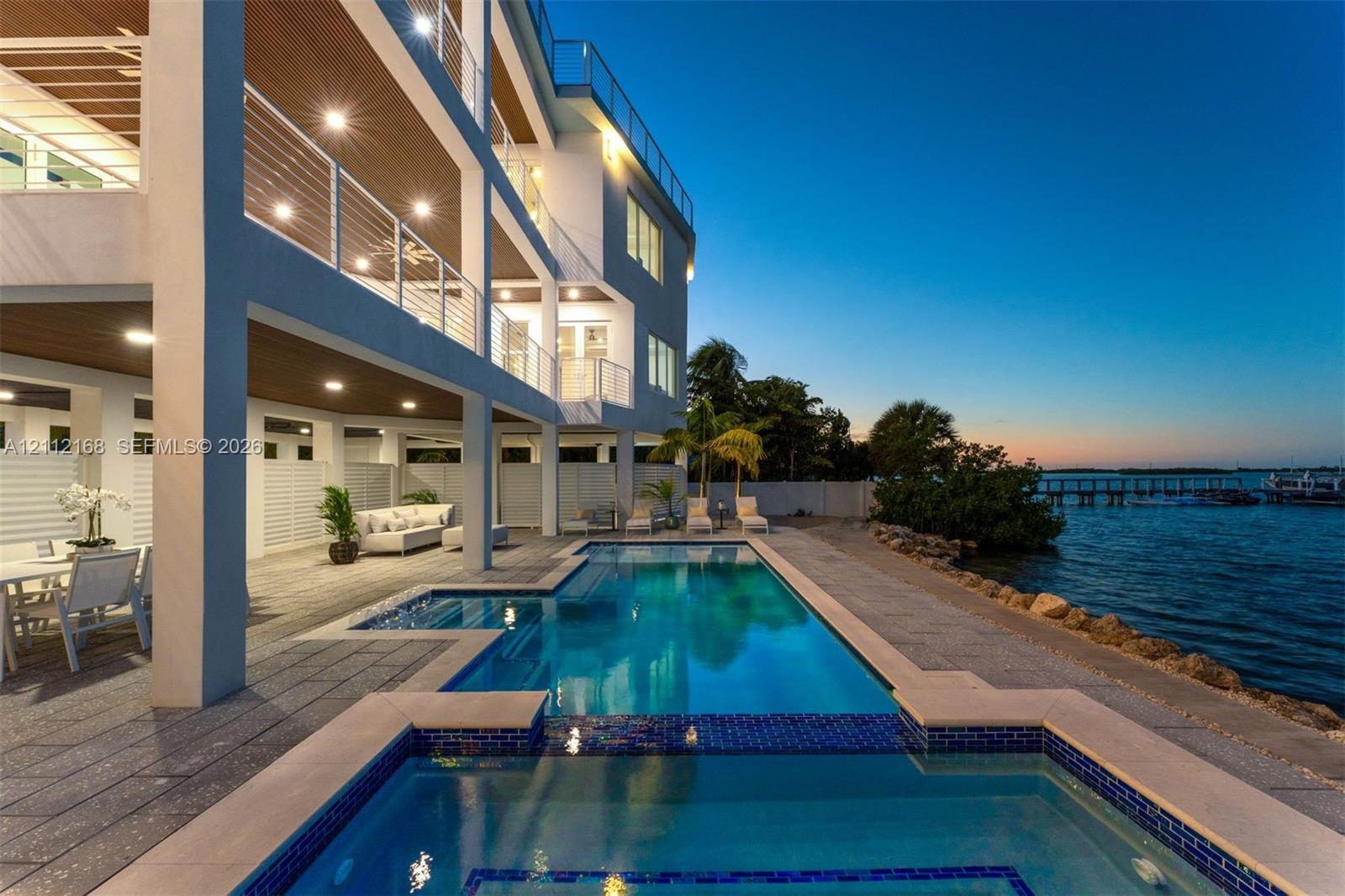 Waterfront multi-level residence featuring a custom swimming pool with integrated spa, expansive covered lanais, metal cable railings, and textured stone-finish decking