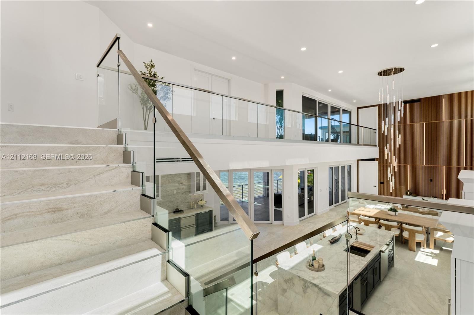 Open-concept interior featuring stone-finish flooring, a glass and metal staircase railing, a multi-level wood-panel wall feature, a stone-topped kitchen island, and multiple glass door systems