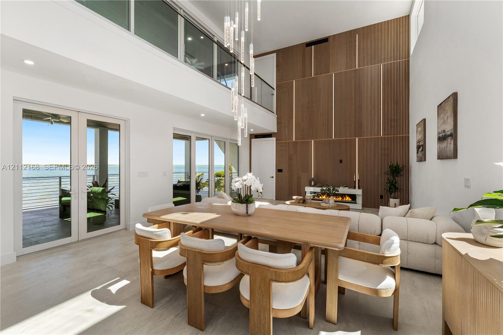 Double-height living area featuring floor-to-ceiling wood paneled accent wall with integrated linear lighting, glass-railed mezzanine, contemporary multi-pendant light fixture, and multiple sets of glass panel doors