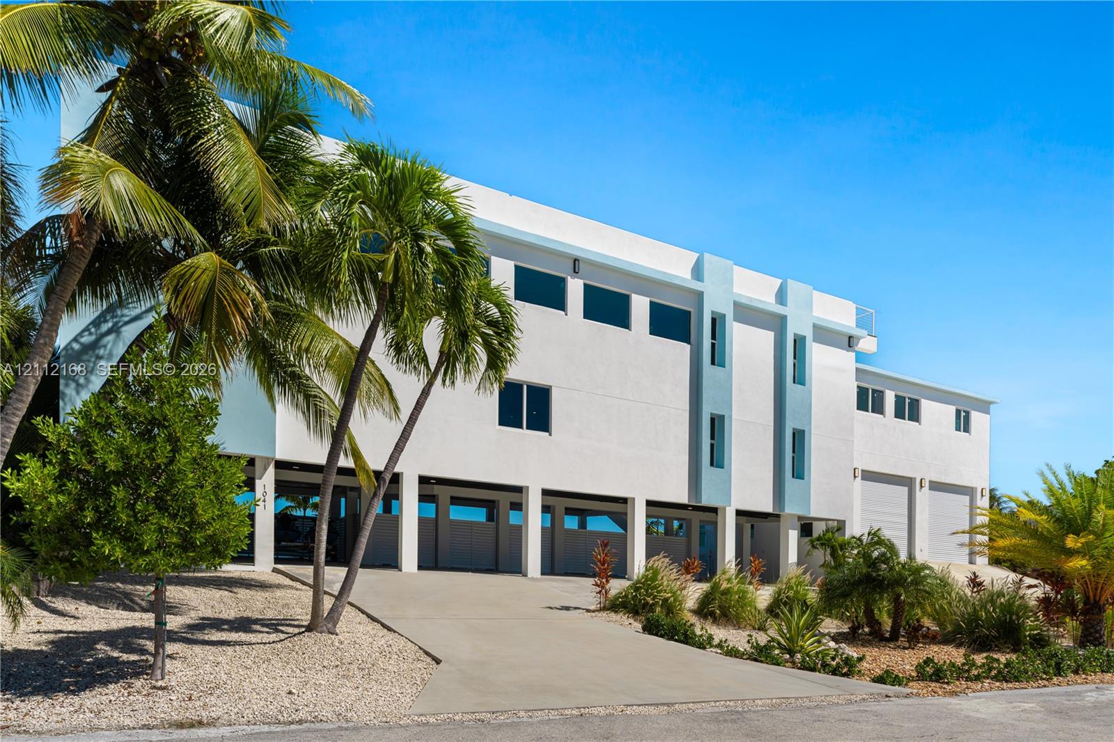 Contemporary multi-level exterior featuring white stucco finish, elevated structural design, integrated garage bays, and mature palm trees