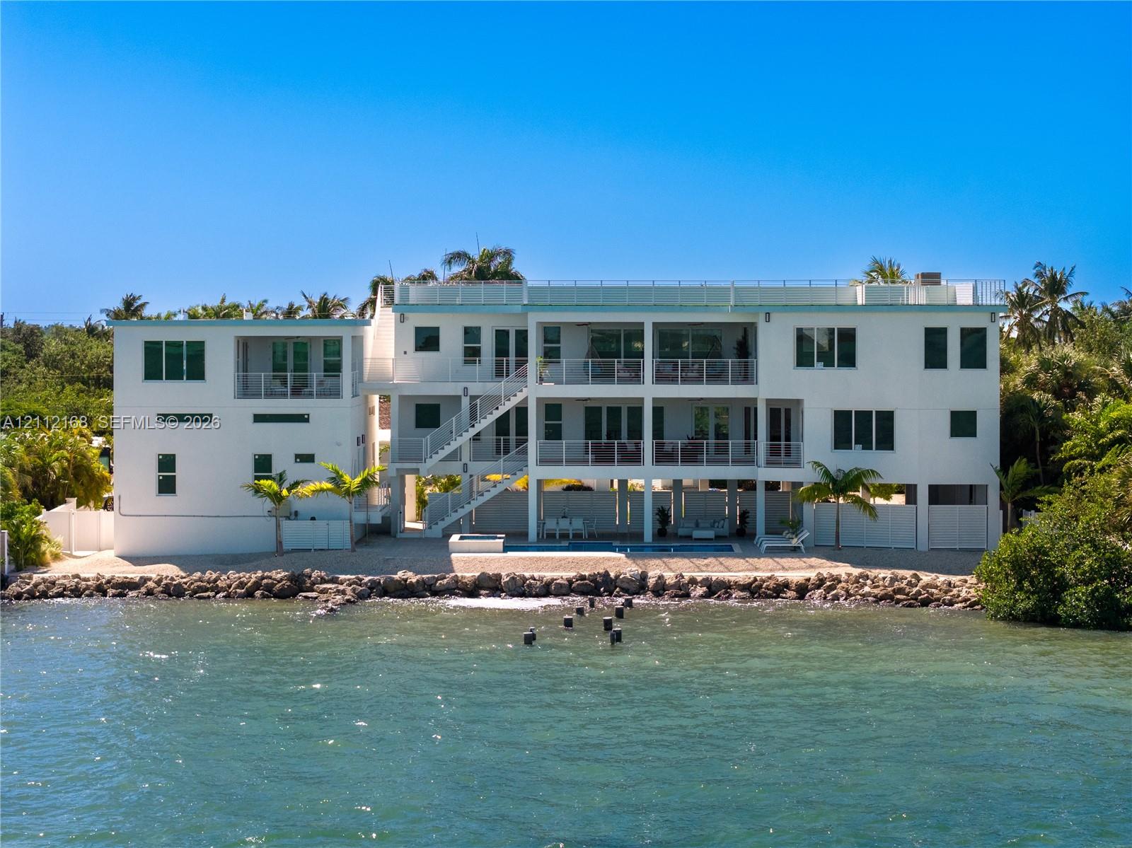 Multi-level waterfront structure featuring a white stucco exterior, tiered balconies with metal railings, an integrated ground-level swimming pool, and stone bulkhead shoreline reinforcement
