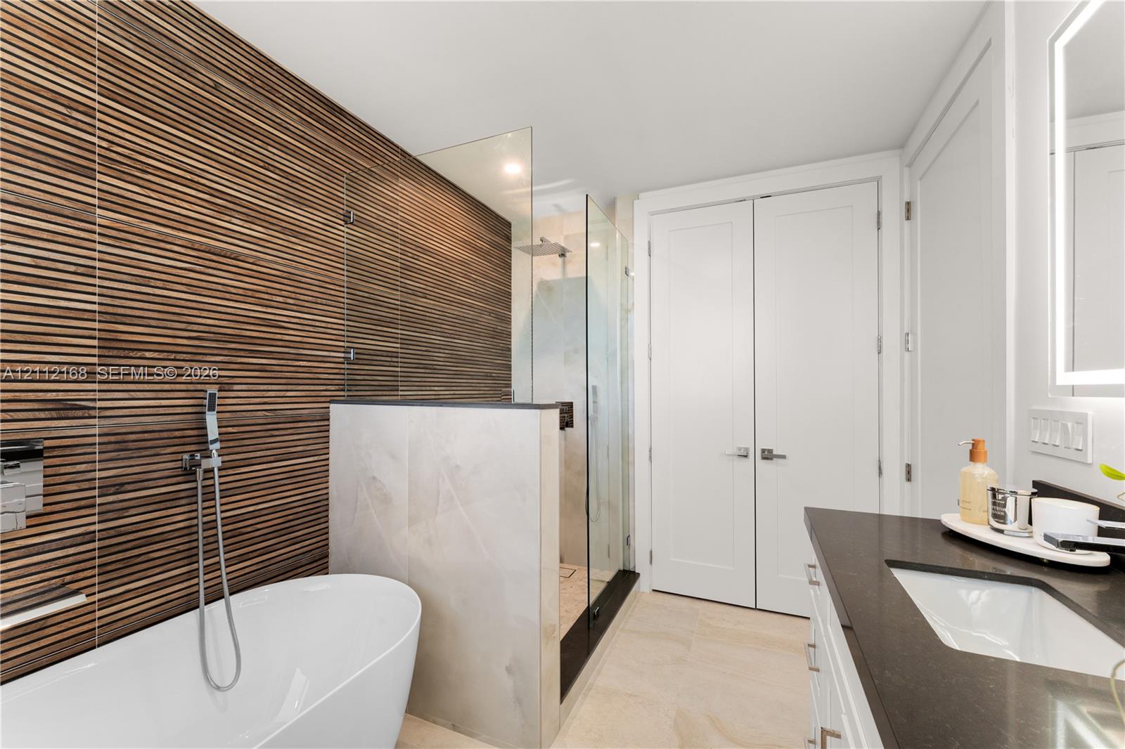 Bathroom featuring a textured wood-slat feature wall, freestanding soaking tub, walk-in glass-enclosed shower, dark countertop with an integrated basin, and double-door storage