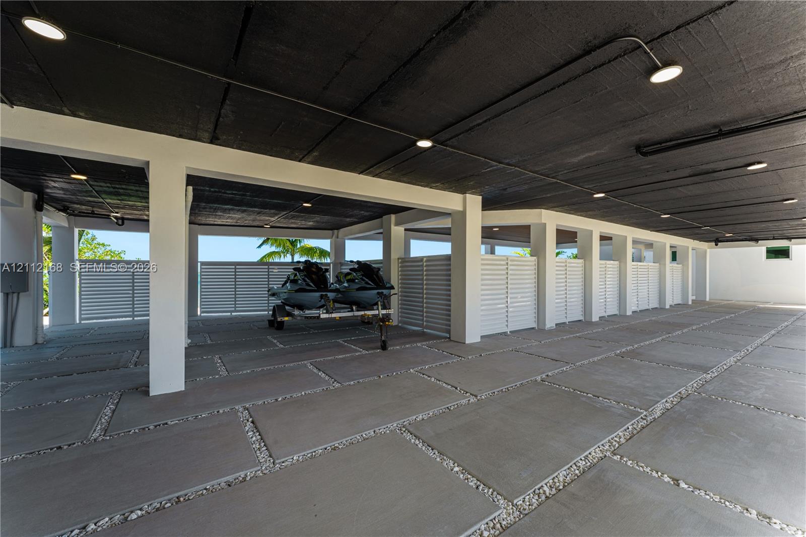 Covered parking area featuring large-format concrete pavers with stone aggregate borders, structural support pillars, white privacy screening, and recessed lighting