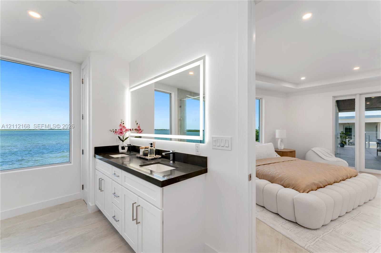 Bathroom vanity featuring white cabinetry, a dark countertop, integrated dual sinks, and an illuminated rectangular mirror, complemented by water-front exterior exposure