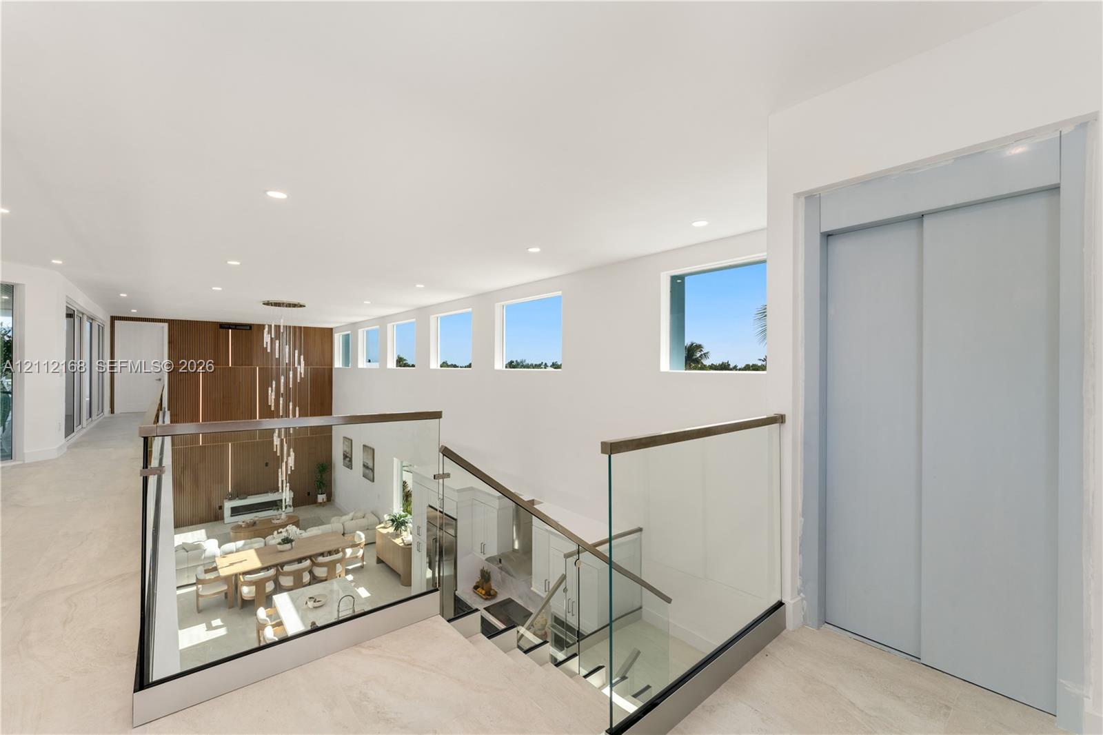 Open landing featuring large-format tile flooring, a glass-panel railing with a wood-finish handrail, a private elevator, recessed ceiling lighting, and a series of high-placement fixed windows