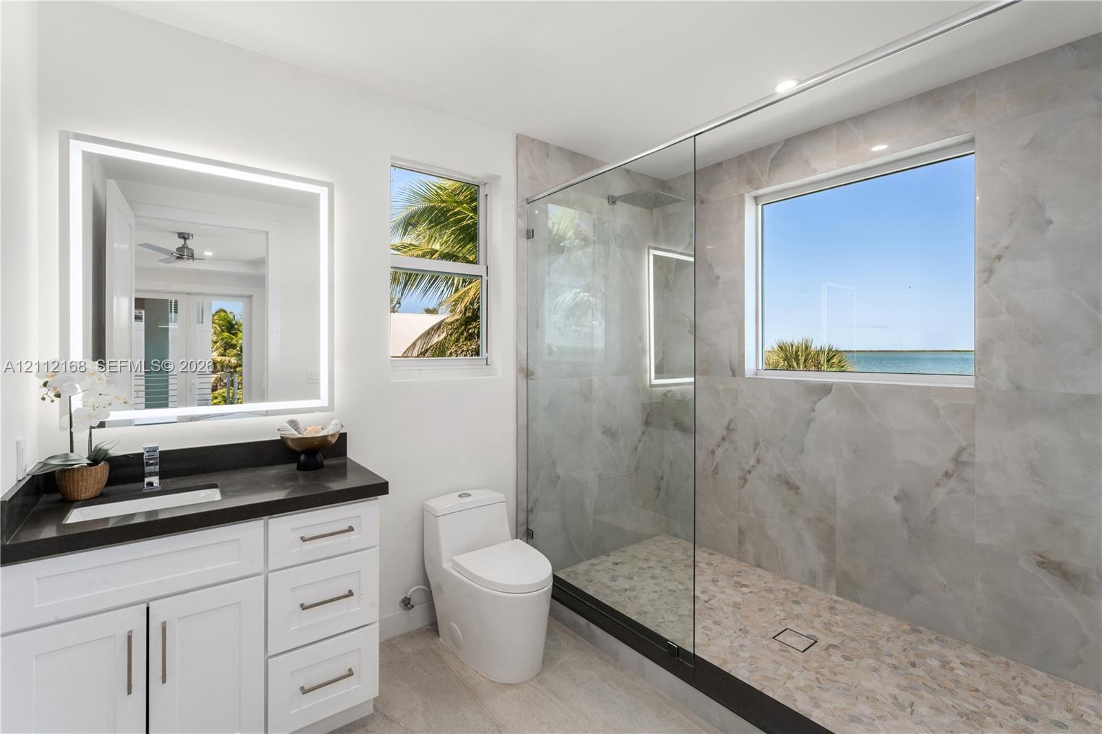 Bathroom featuring a white vanity with a dark countertop, an illuminated mirror, a glass-enclosed shower with large-format tile surround, and an integrated window