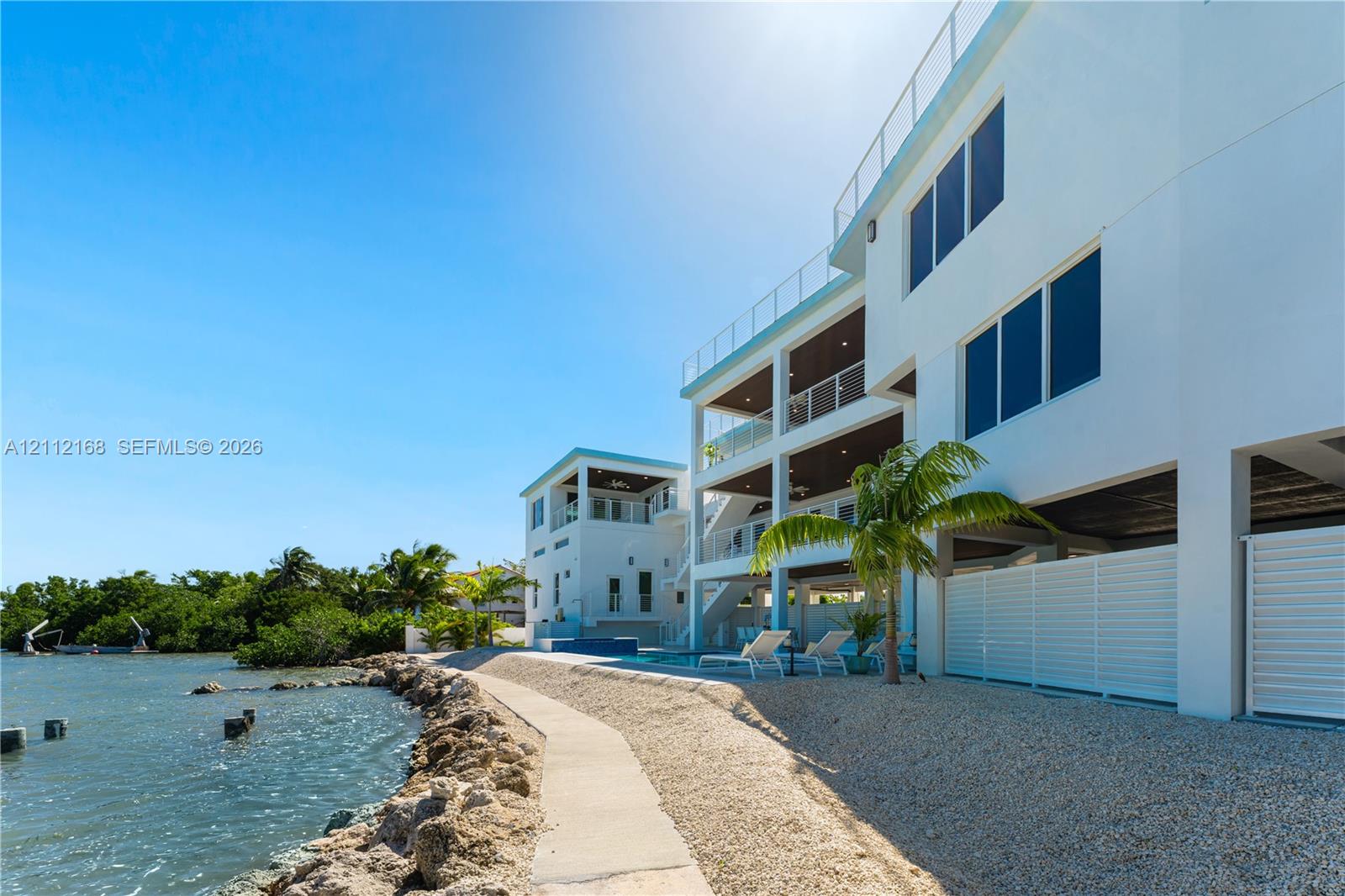 Multi-level waterfront exterior featuring white stucco siding, glass-panel railings, covered balconies, an inground swimming pool, and stone-lined shoreline