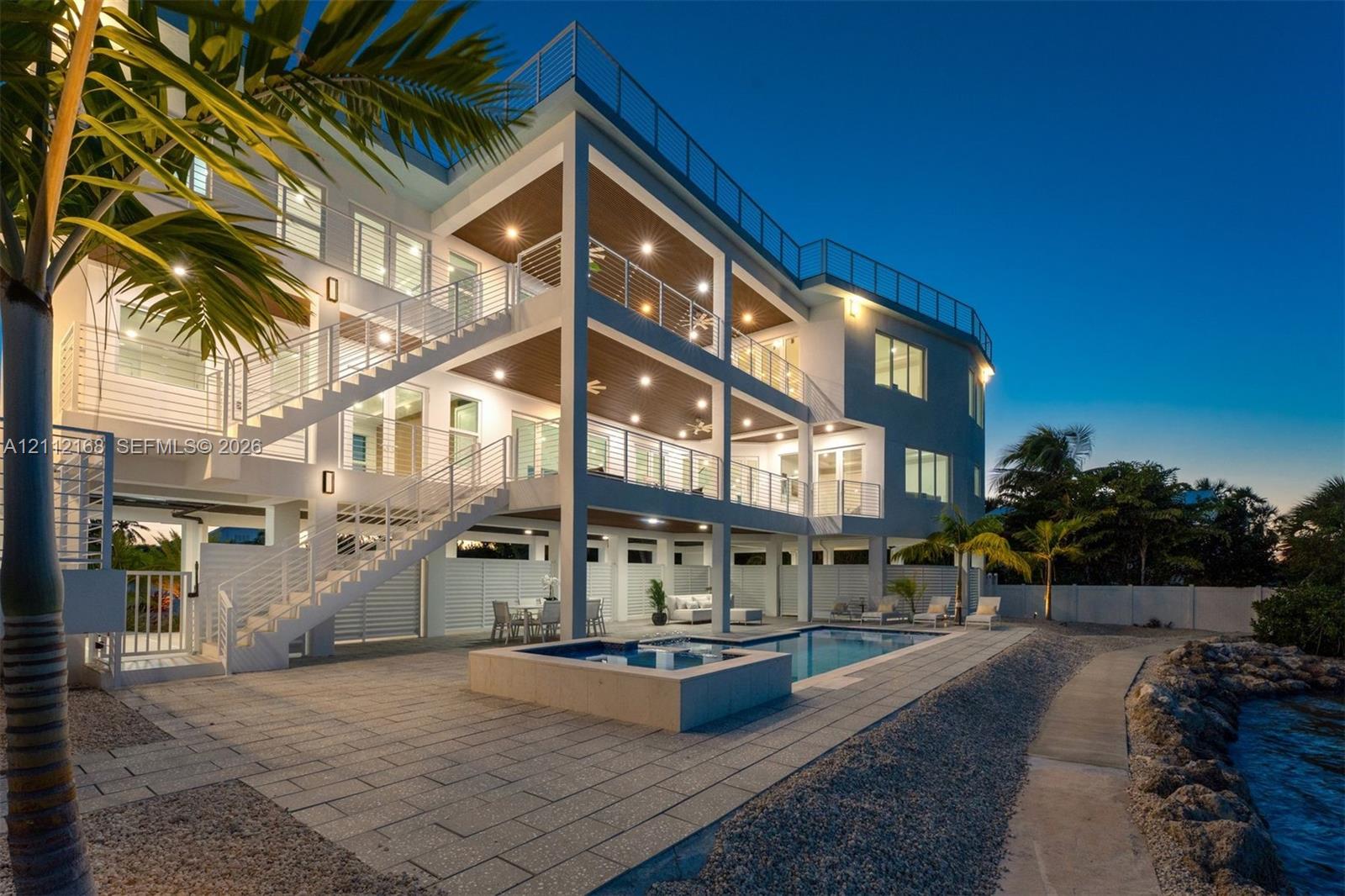 Multi-level exterior featuring stacked covered balconies, a rooftop terrace with horizontal cable railings, a built-in swimming pool and spa, and stone-paver hardscaping