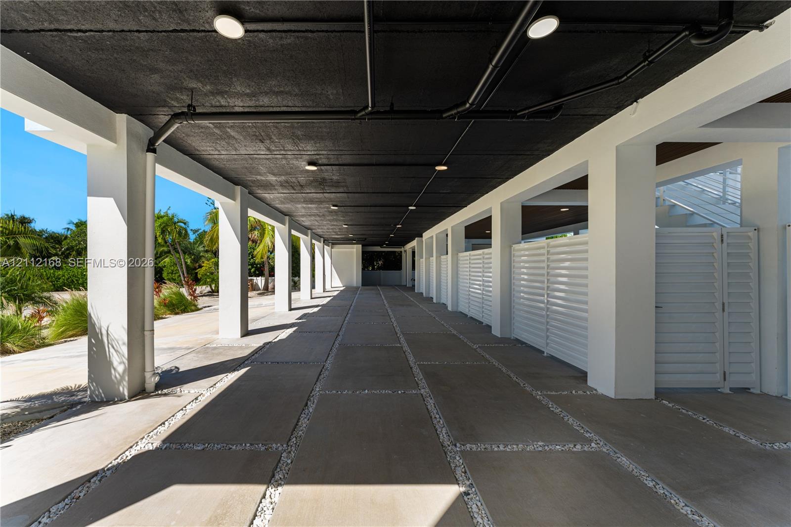 Covered ground-level parking area featuring concrete pavers, white louvered privacy screens, recessed lighting, and exposed ceiling structure